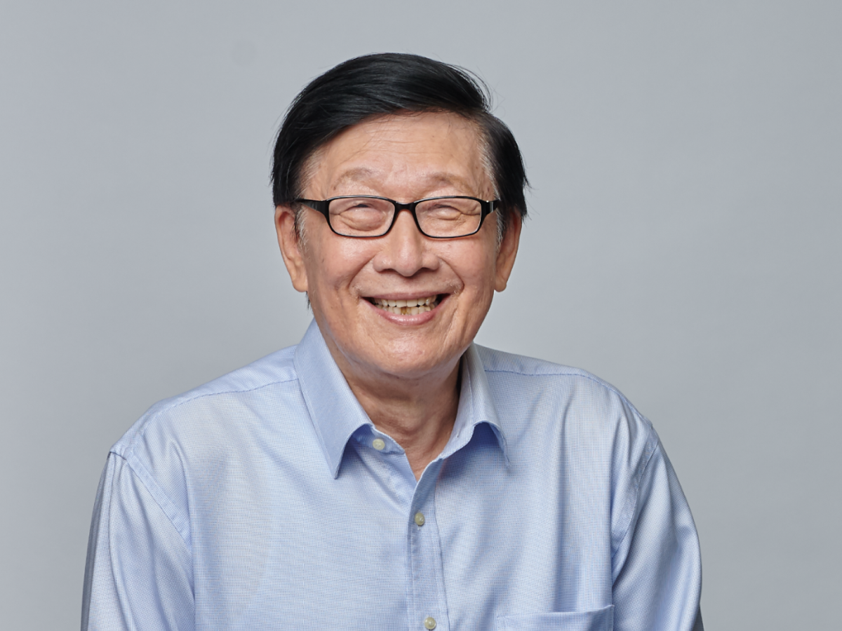 Mr Lim Bok Ngam in glasses wearing a light blue shirt smiling with hands clasped against a plain grey background